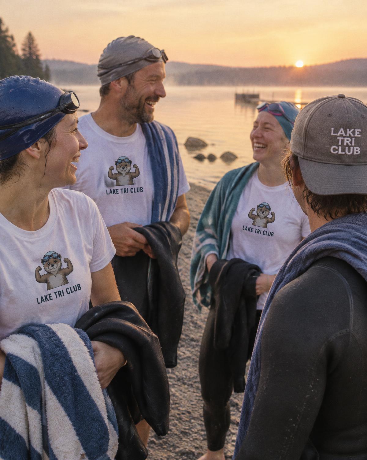 Swimmers in white Lake Tri Club tees and a grey stitched cap laughing on the shore with wetsuits.