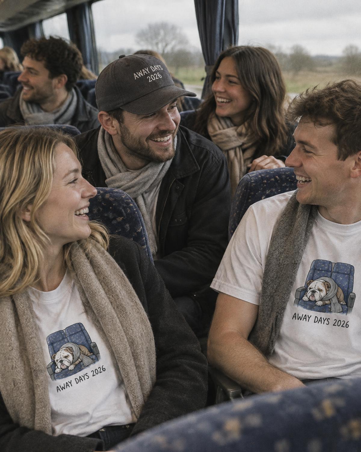 Friends in white Away Days 2026 tees and a black stitched cap laughing in their seats on the coach.