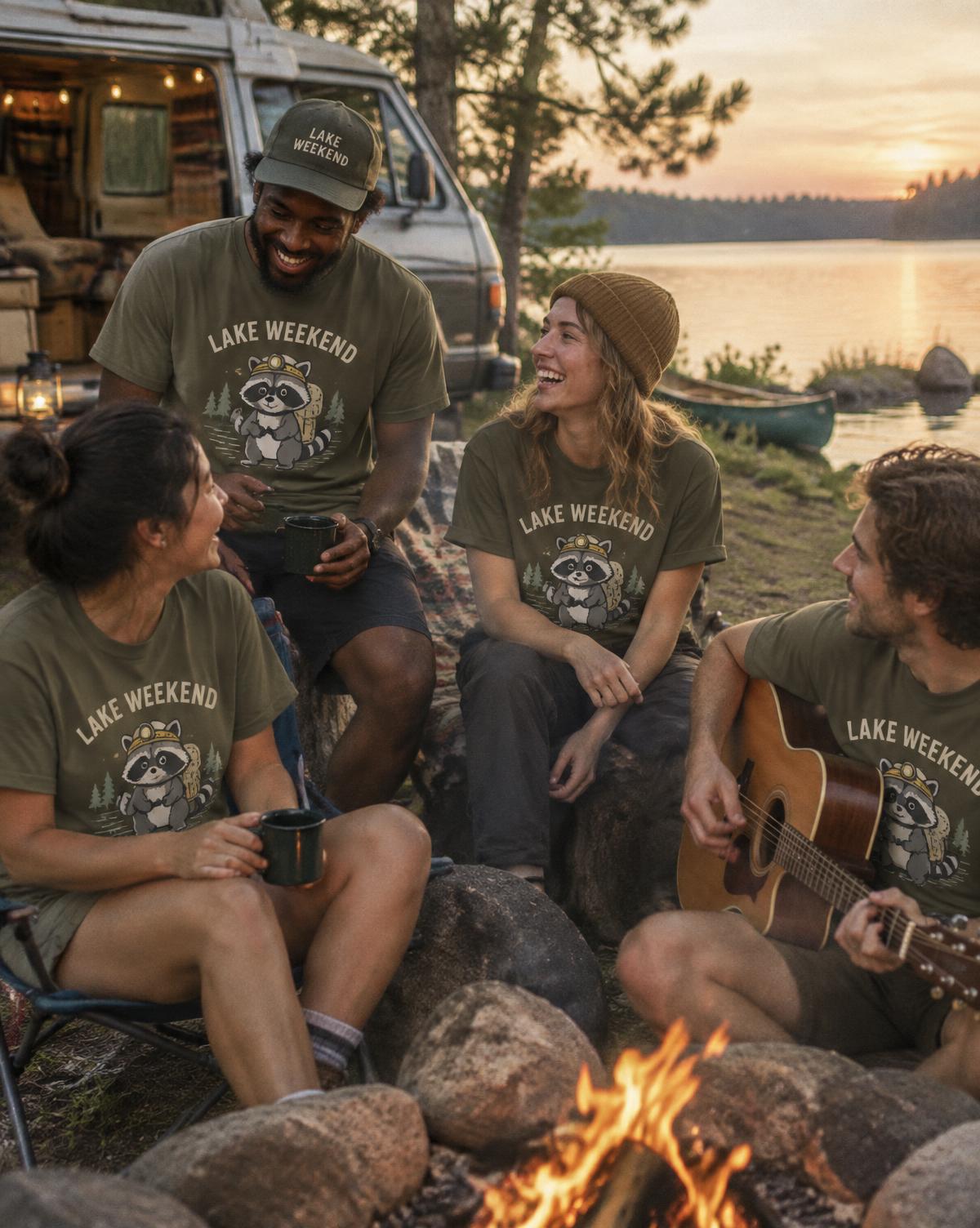 Friends in olive Lake Weekend tees with a raccoon print laugh around a campfire, one playing guitar.