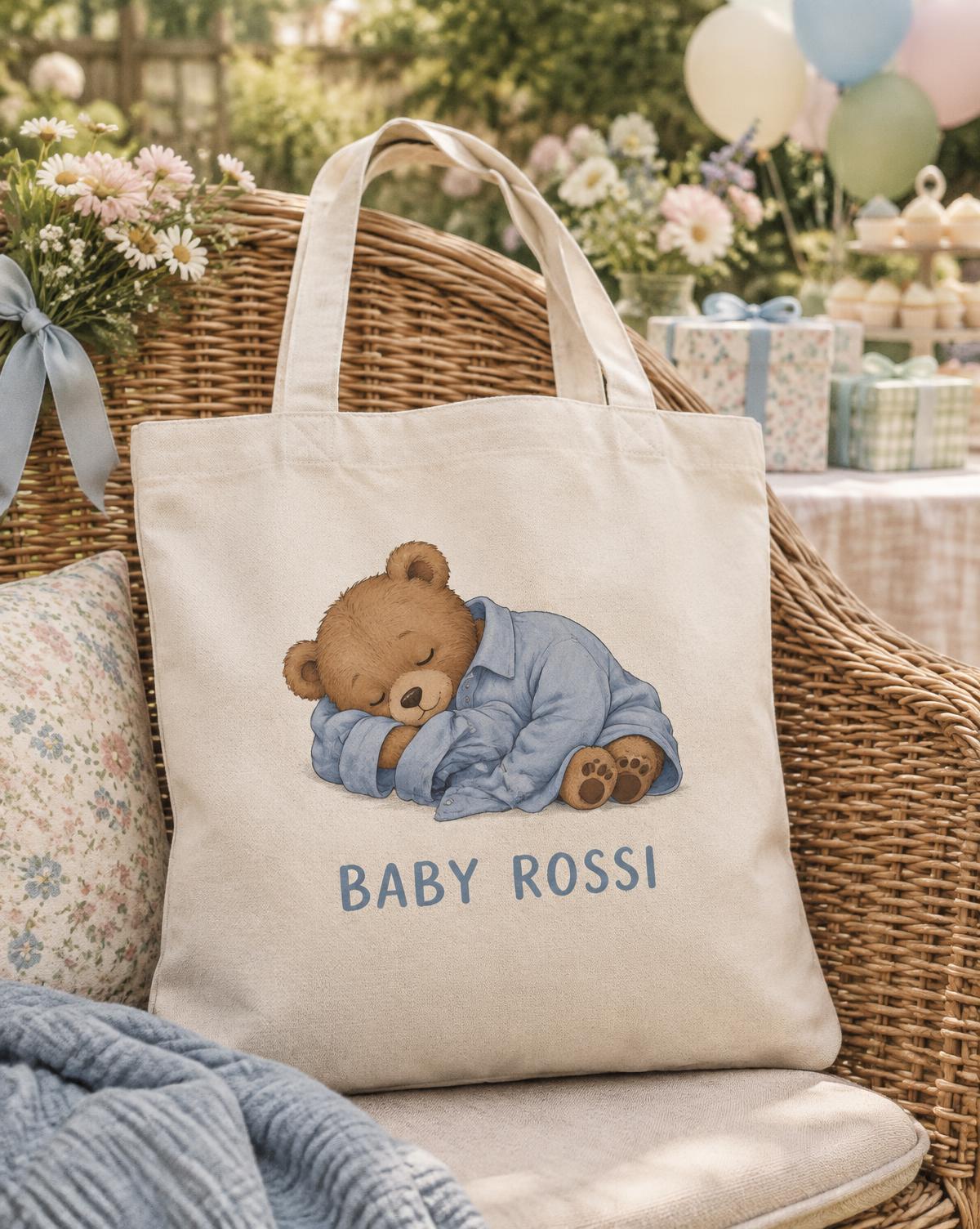 Natural tote printed with a bear asleep in a blue shirt and Baby Rossi lettering, resting on a wicker garden chair.