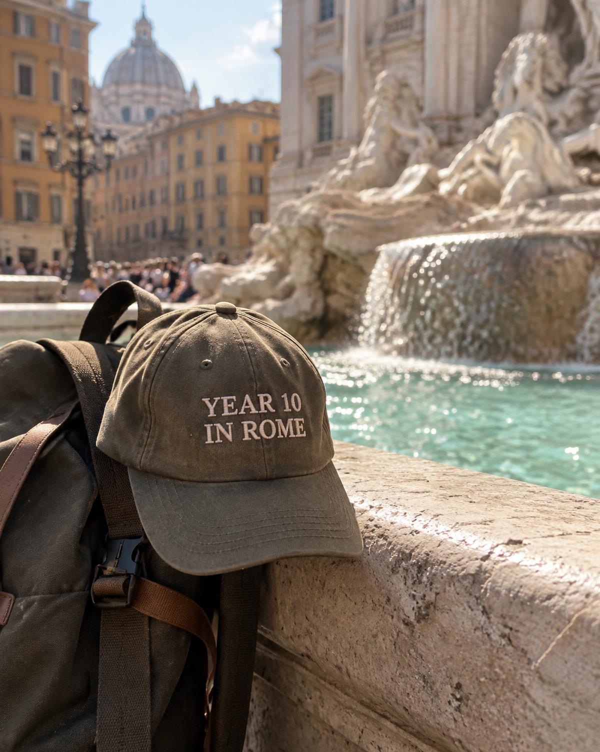 Olive cap with Year 10 In Rome stitched in cream, hanging on a backpack at the edge of a fountain.