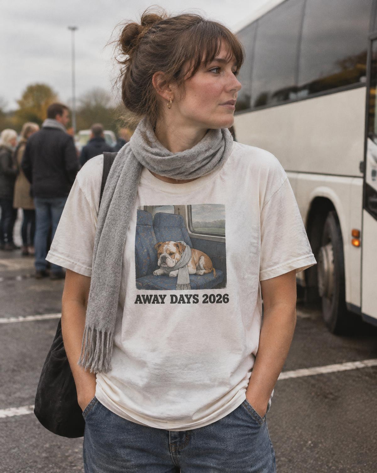 Woman in a white Away Days 2026 tee with a bulldog asleep on a bus seat, waiting by the coaches.