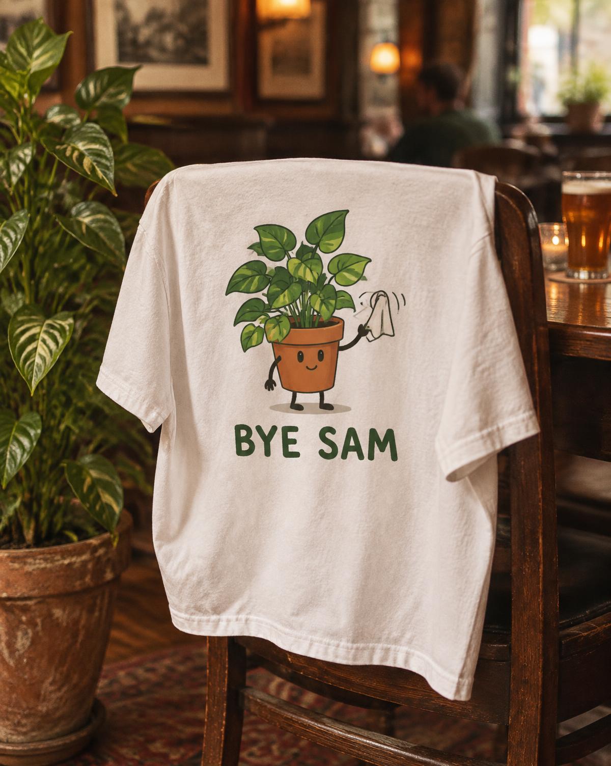 White Bye Sam tee with a potted plant waving a handkerchief, draped over a chair in a cosy pub.