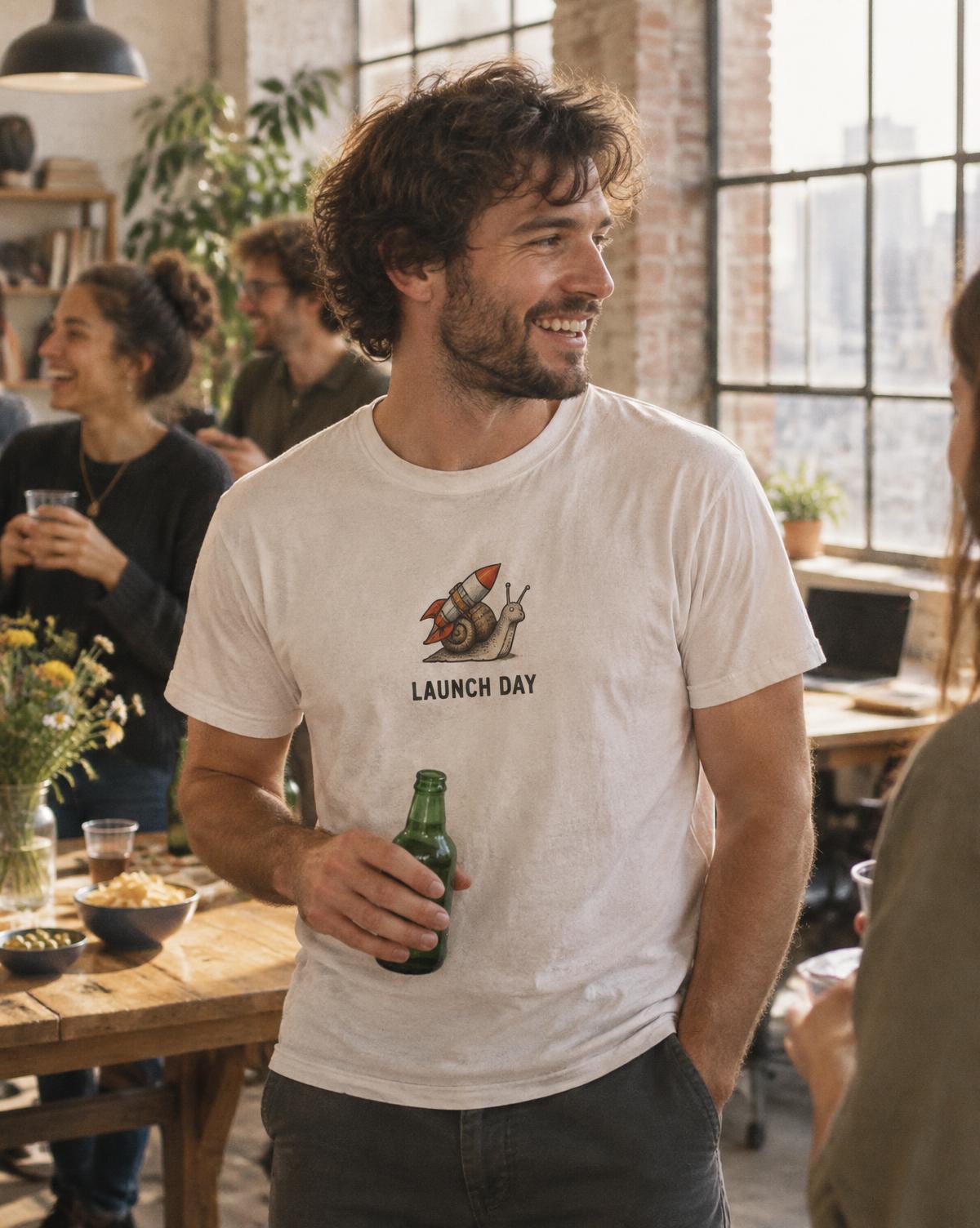 Smiling man in a white Launch Day tee with a rocket snail print holding a beer at an office party.