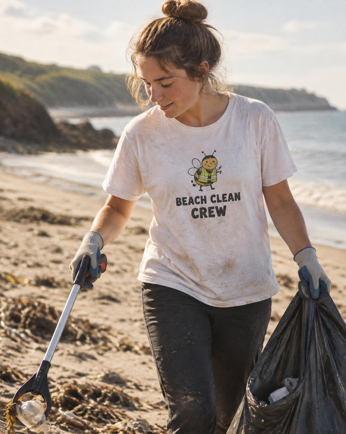 Volunteer in a white Beach Clean Crew tee with a bee print collecting litter with a picker and bin bag.