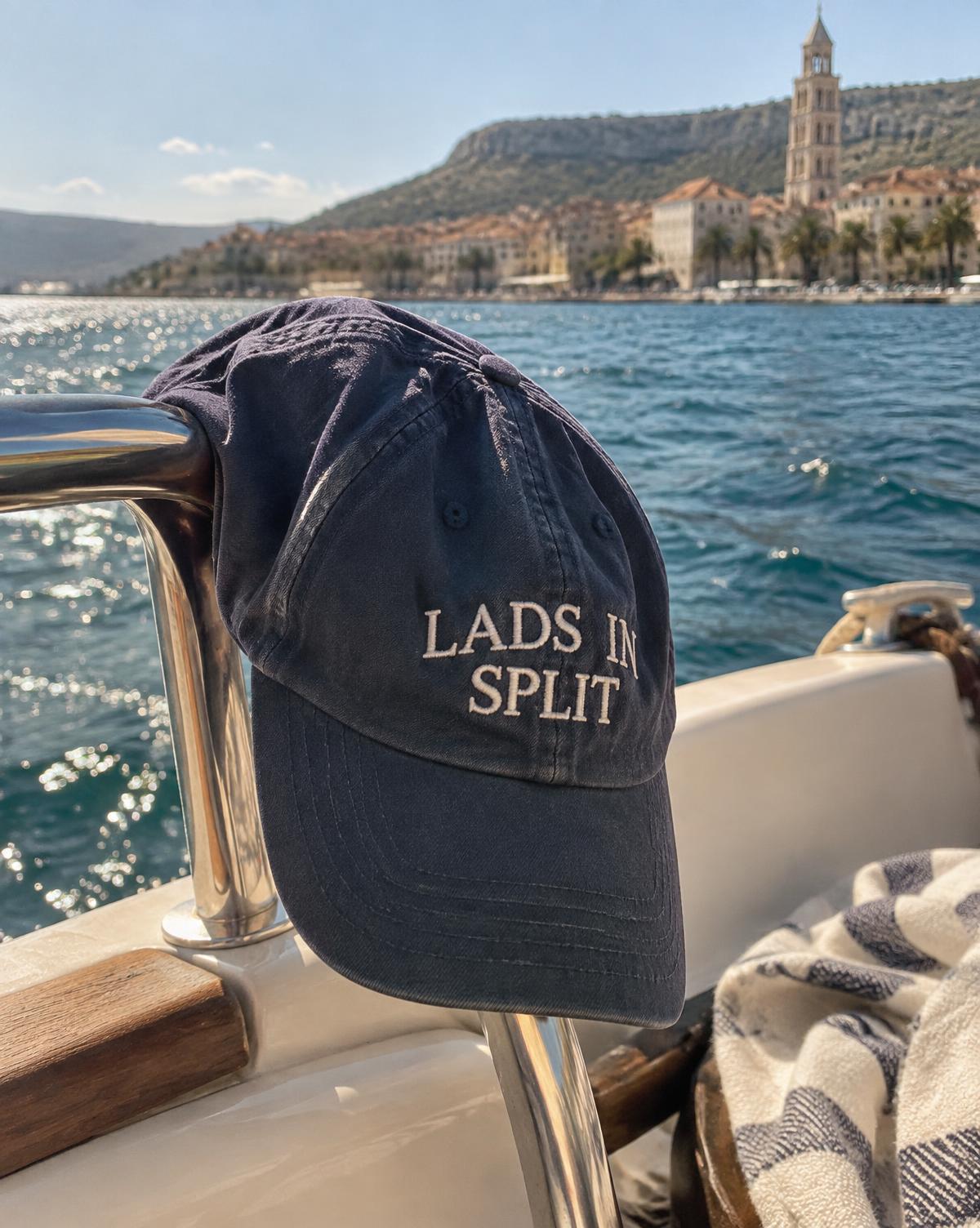 Navy cap with Lads In Split stitched in white, hanging on a boat rail with the old town of Split behind.