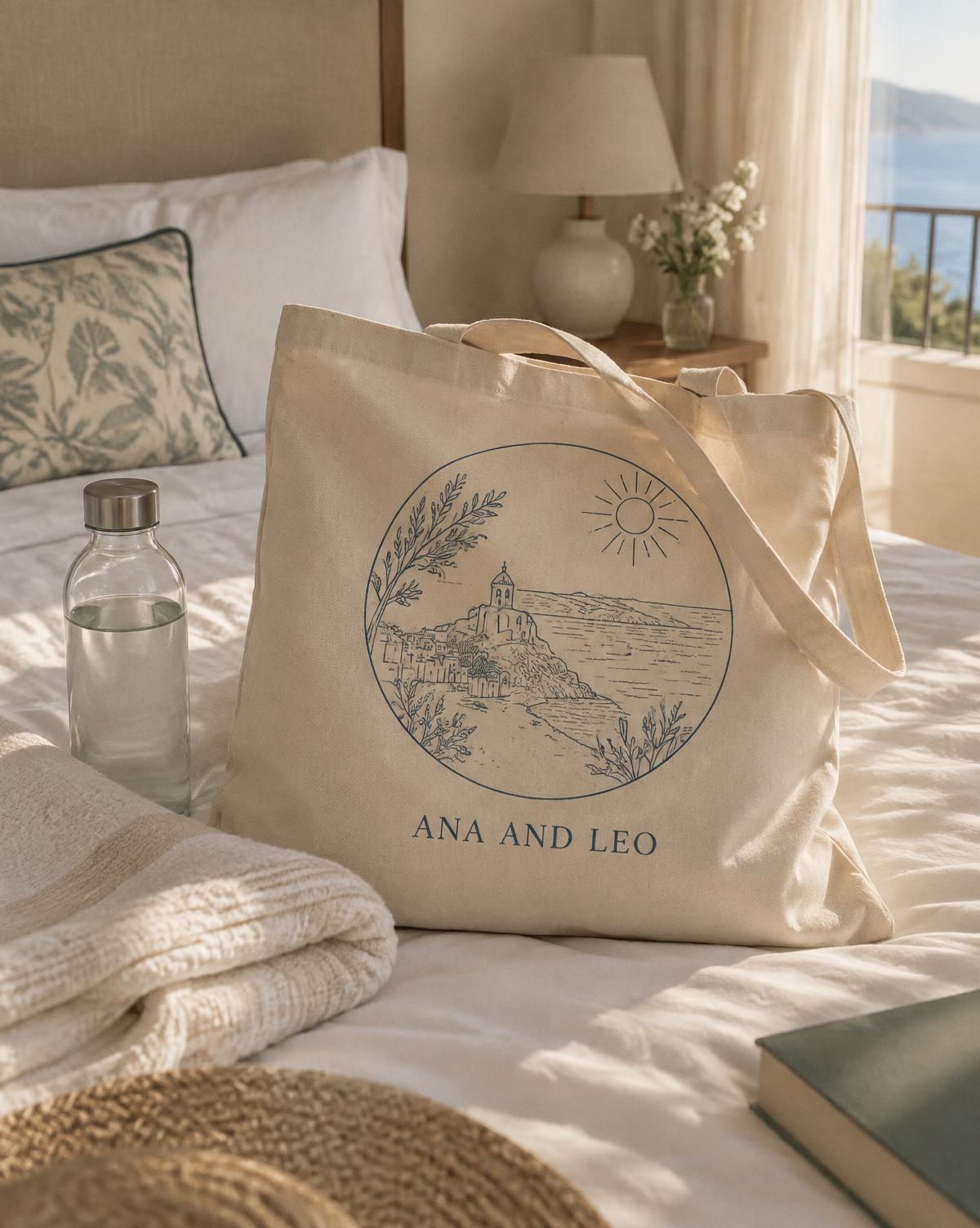 Natural Ana And Leo tote with a seaside village sketch, laid on a hotel bed beside a bottle of water.