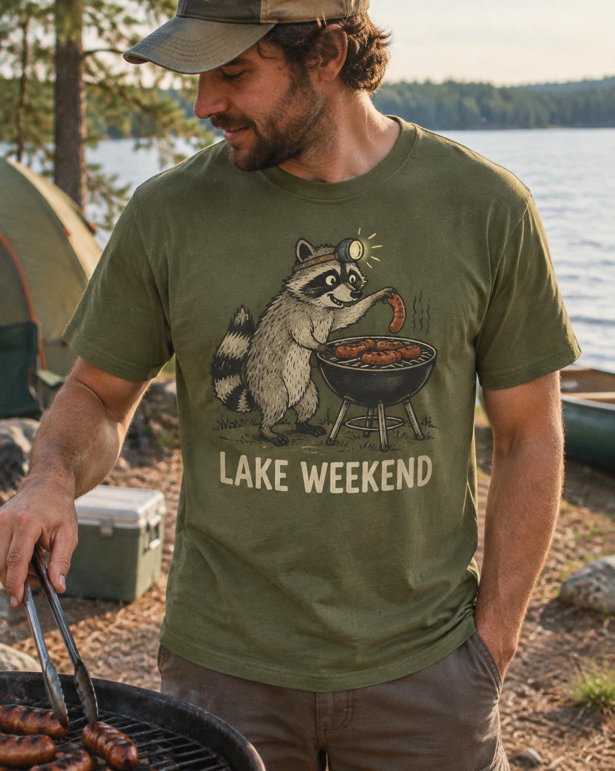 Man grilling in an olive Lake Weekend tee printed with a raccoon in a headlamp stealing a sausage.
