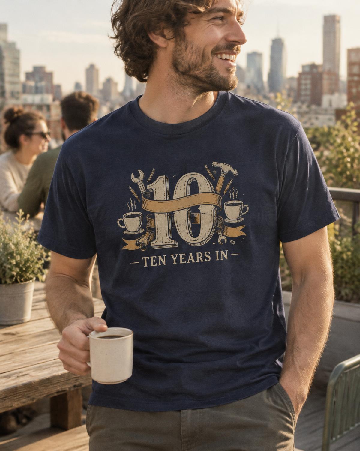 Smiling man in a navy Ten Years In tee with a 10, ribbon, tools and coffee cups print on a rooftop.