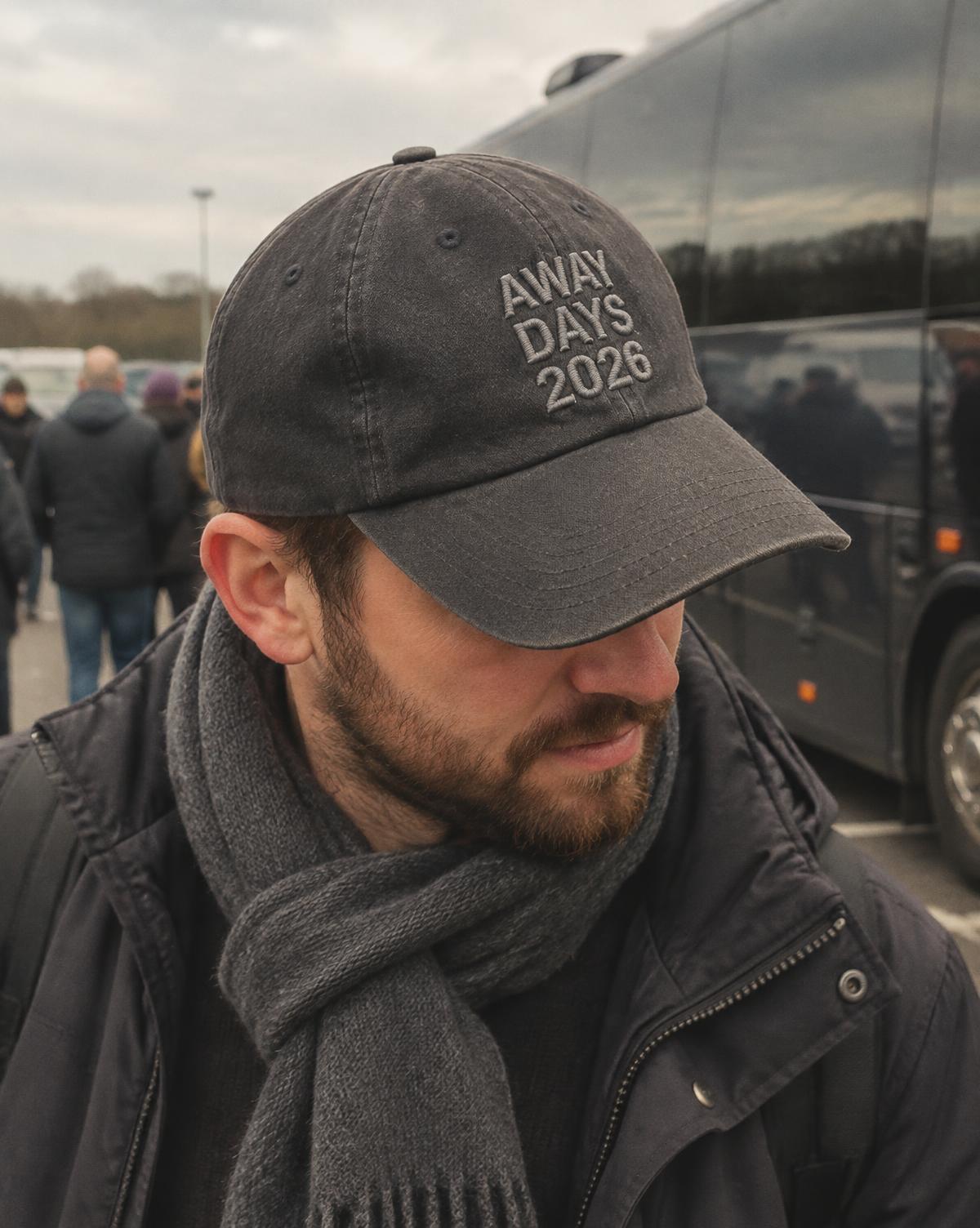 Fan in a charcoal cap with Away Days 2026 stitched in grey and a wool scarf in a coach car park.