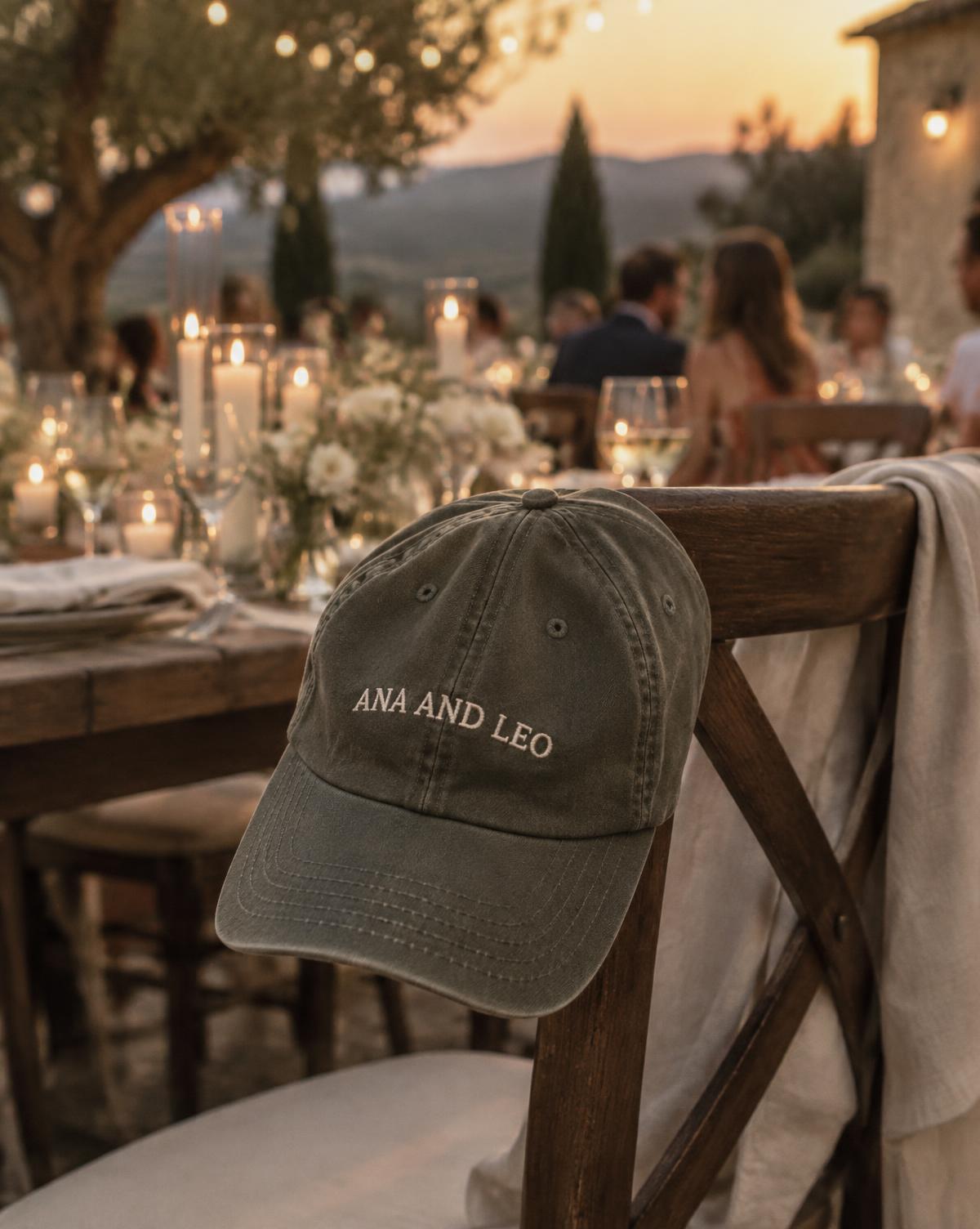 Olive cap with Ana And Leo stitched in cream, hanging on a chair at a candlelit outdoor wedding dinner.