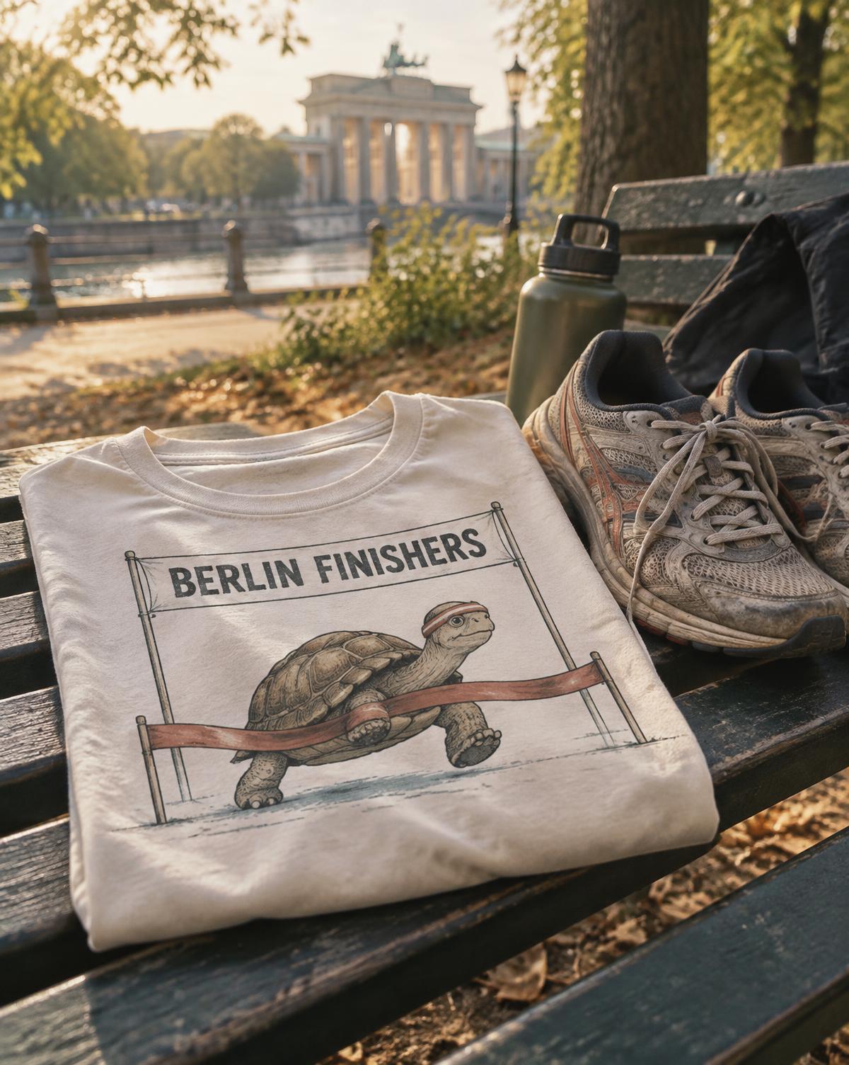 Folded cream Berlin Finishers tee with a tortoise at the finish tape, on a park bench by muddy trainers.