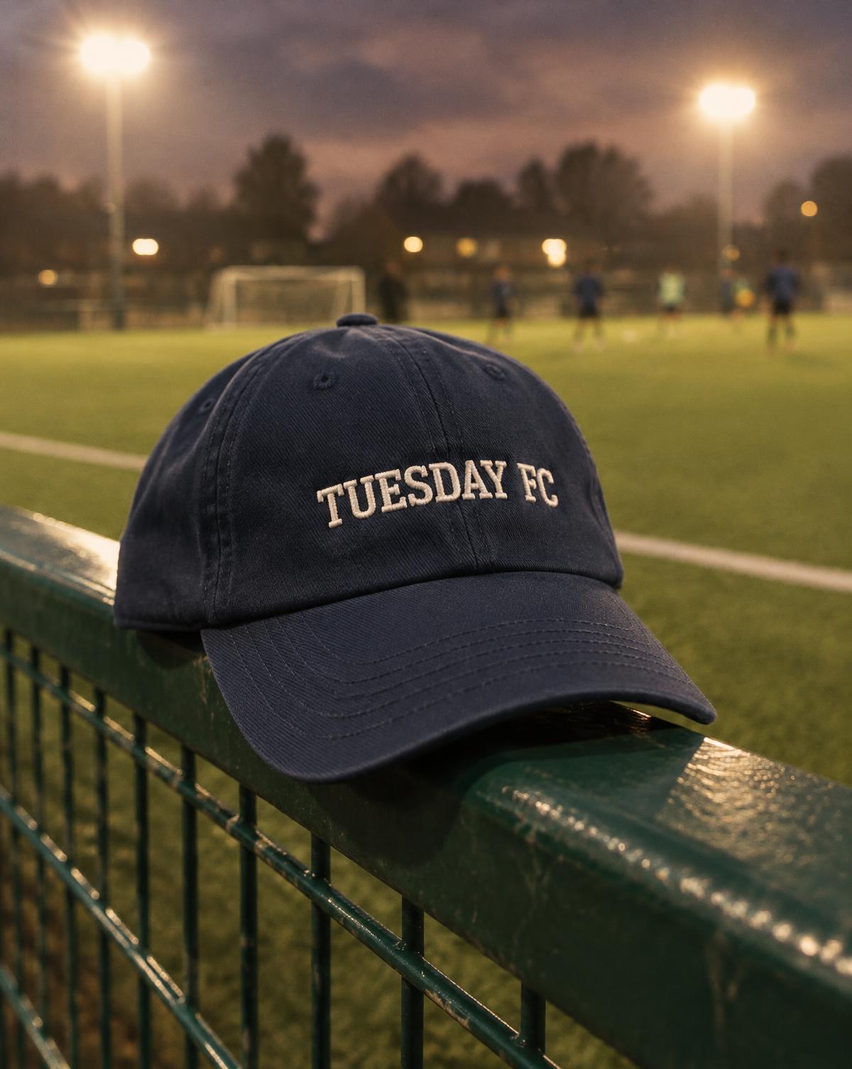 Navy cap with Tuesday FC stitched in cream, resting on a pitch-side railing under evening floodlights.