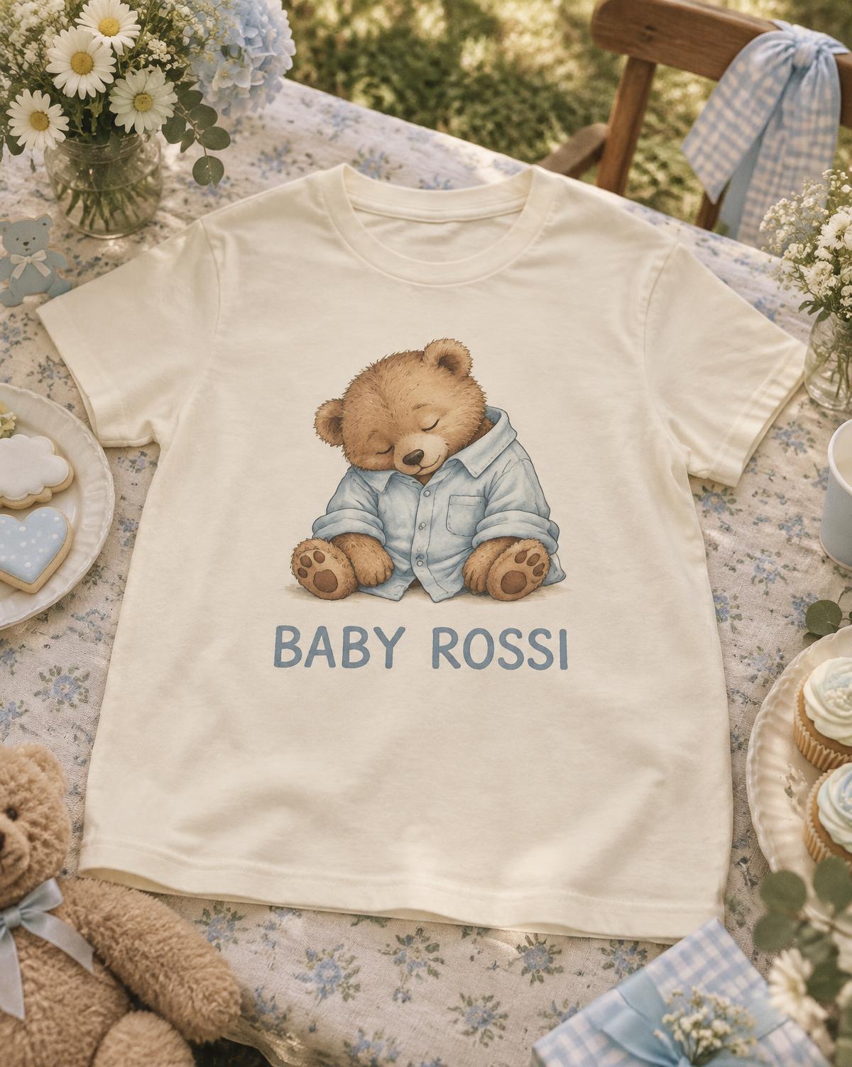 Cream Baby Rossi tee with a sleepy bear in a blue shirt laid flat on a floral table with cookies and cupcakes.