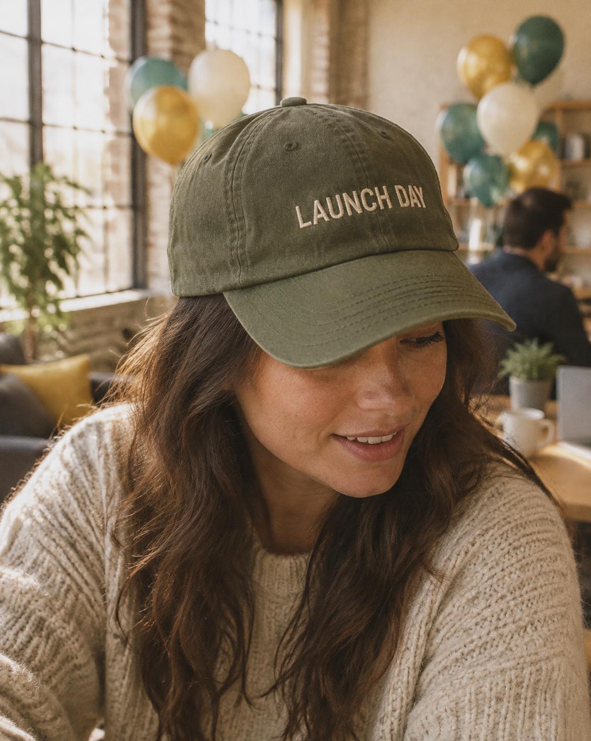 Woman in an olive cap with Launch Day stitched in cream, smiling down at a laptop with balloons behind.