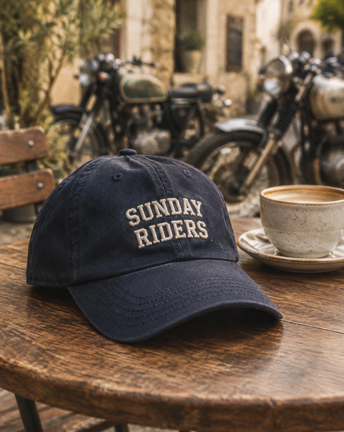Navy cap with Sunday Riders in raised cream stitching on a cafe table, vintage motorbikes behind.
