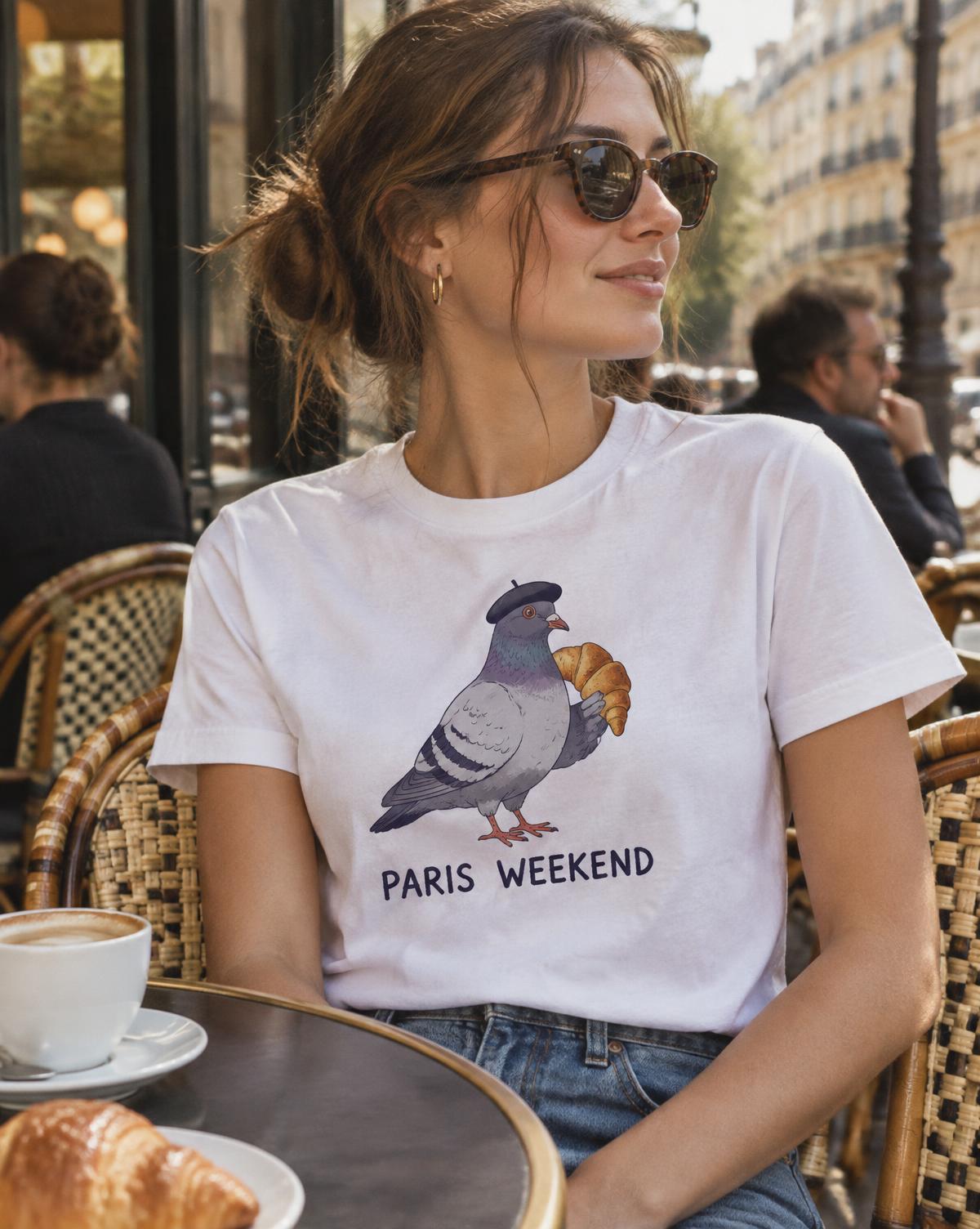 Woman in a white Paris Weekend tee with a pigeon in a beret holding a croissant at a cafe table.
