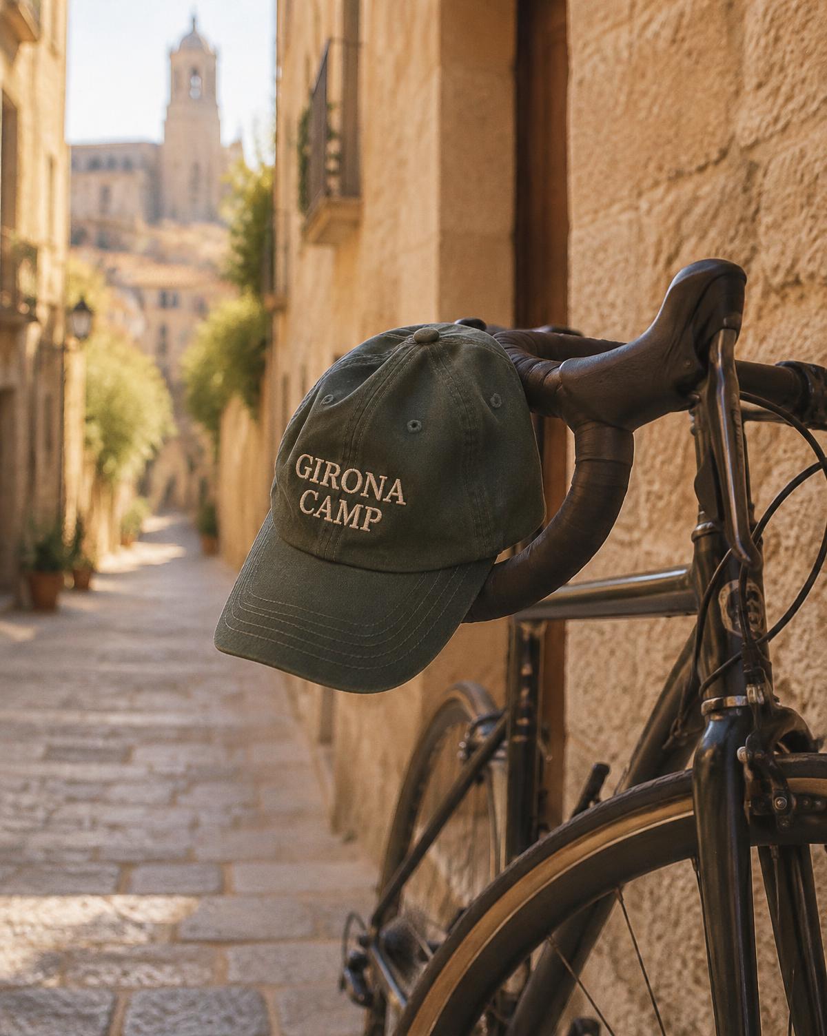 Dark green cap with Girona Camp stitched in cream, hung on a road bike in a narrow stone lane.