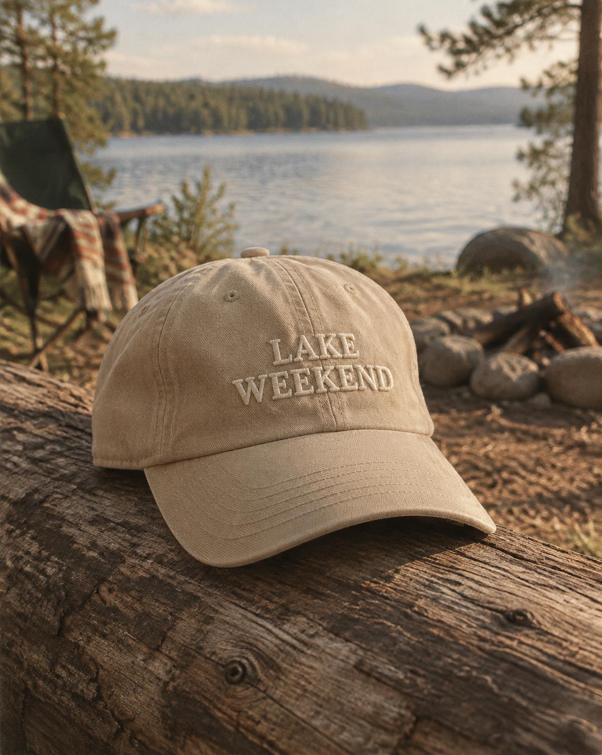 Sand coloured cap with Lake Weekend in raised stitching, lying on a log by a lakeside campfire.