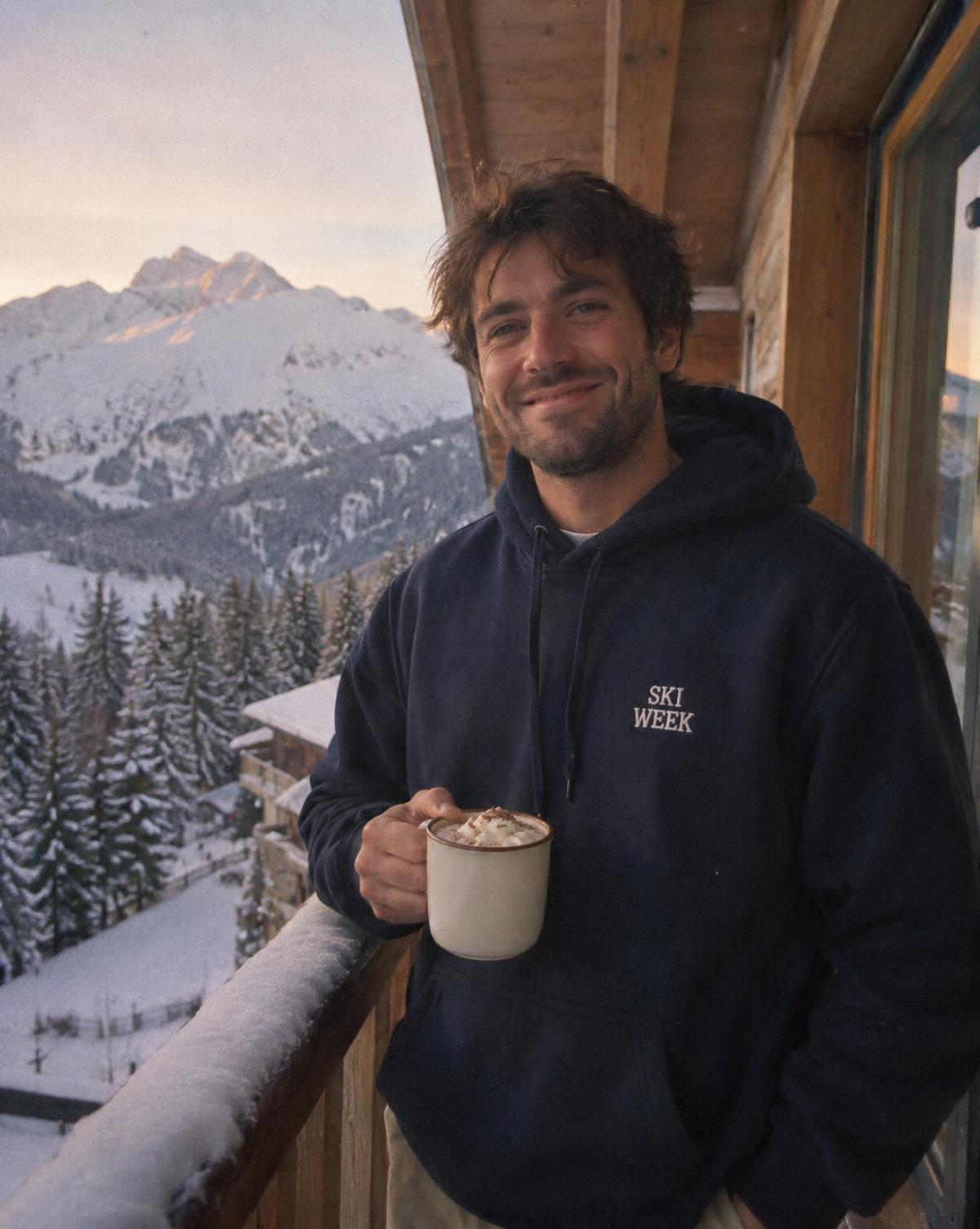 Man on a snowy chalet balcony in a navy hoodie with white embroidered Ski Week patch